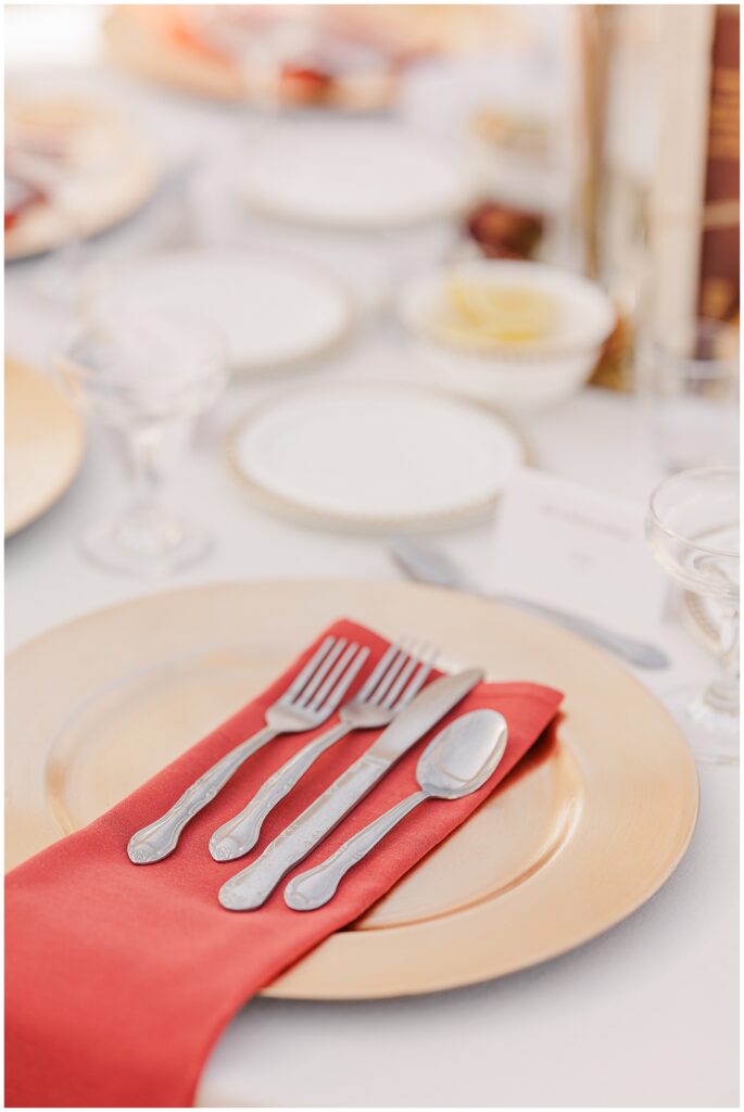 A table setting at the Colonial Hotel wedding reception in Gardner, MA, features a gold charger plate topped with a red napkin and silver cutlery, including two forks, a knife, and a spoon, neatly arranged on top. In the background, additional white plates, glassware, and decor elements complete the elegant fall-inspired setup.