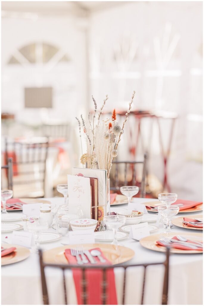 A wedding reception table centerpiece at the Colonial Hotel with a fall-inspired arrangement of dried flowers, including soft, neutral tones and pops of orange and red. The table is set with gold-rimmed plates, glassware, and red napkins, with other tables and chairs visible in the background.