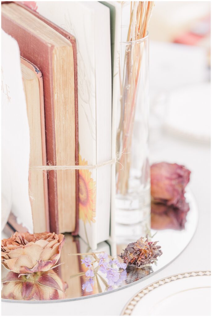 A close-up of a vintage book-themed wedding centerpiece, featuring several books tied together with twine alongside dried flowers, all arranged on a reflective surface. The books’ spines, aged and colorful, add a nostalgic touch to the table decor at the Colonial Hotel wedding.