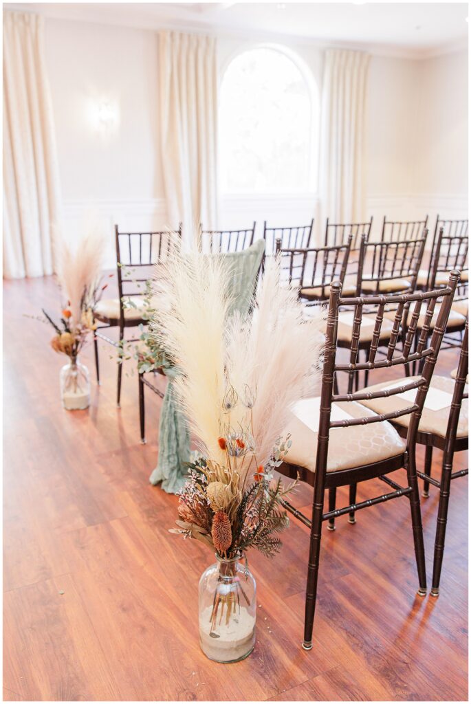 An indoor wedding ceremony setup at the Colonial Hotel with rows of dark wooden chairs arranged on a polished wood floor. Large glass vases filled with pampas grass, eucalyptus, and other dried florals line the aisle, adding a natural, fall-inspired decor to the intimate ceremony space.