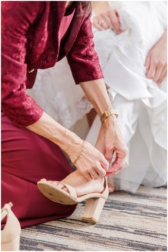 A close-up of a woman, possibly the mother of the bride, helping the bride by fastening a beige, block-heeled sandal on her foot. The woman is dressed in a deep red lace outfit, and her hands are adorned with a watch and a bracelet, adding a tender, personal touch to the getting-ready moment.