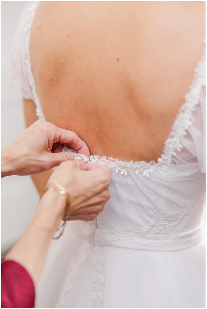 A close-up of the back of a bride’s lace wedding dress as a woman helps fasten the delicate button closure. The intricate lace detail on the dress is visible, along with the woman’s hands as she assists with the finishing touches on the bride’s attire.