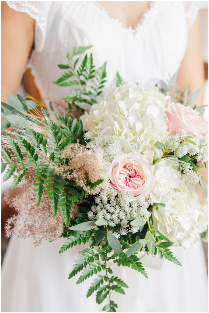 A bride holds a lush bouquet of white hydrangeas, soft pink roses, Queen Anne’s lace, and green ferns. The arrangement has a natural, slightly wild look, adding a fresh, romantic feel to the New England fall wedding at the Colonial Hotel.