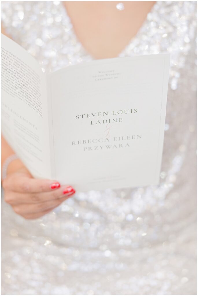 A wedding guest or bridal party member holds a program for the wedding ceremony at the Colonial Hotel in Gardner, MA. The program cover reads “Steven Louis Ladine & Rebecca Eileen Przywara” in elegant text, with the person’s hand and red nails visible against a glittering, silver fabric background.