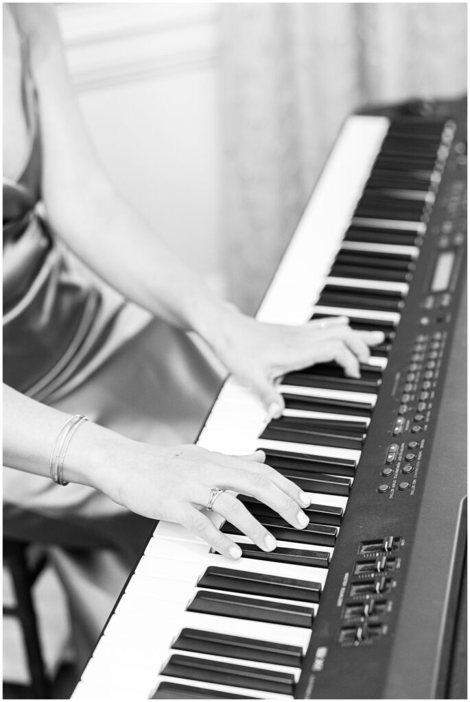 A black-and-white close-up of a woman’s hands playing a keyboard, wearing a ring and bracelet, adding a musical ambiance to the wedding at the Colonial Hotel. The keys and controls on the keyboard are clearly visible, capturing the elegance and focus of the live performance.