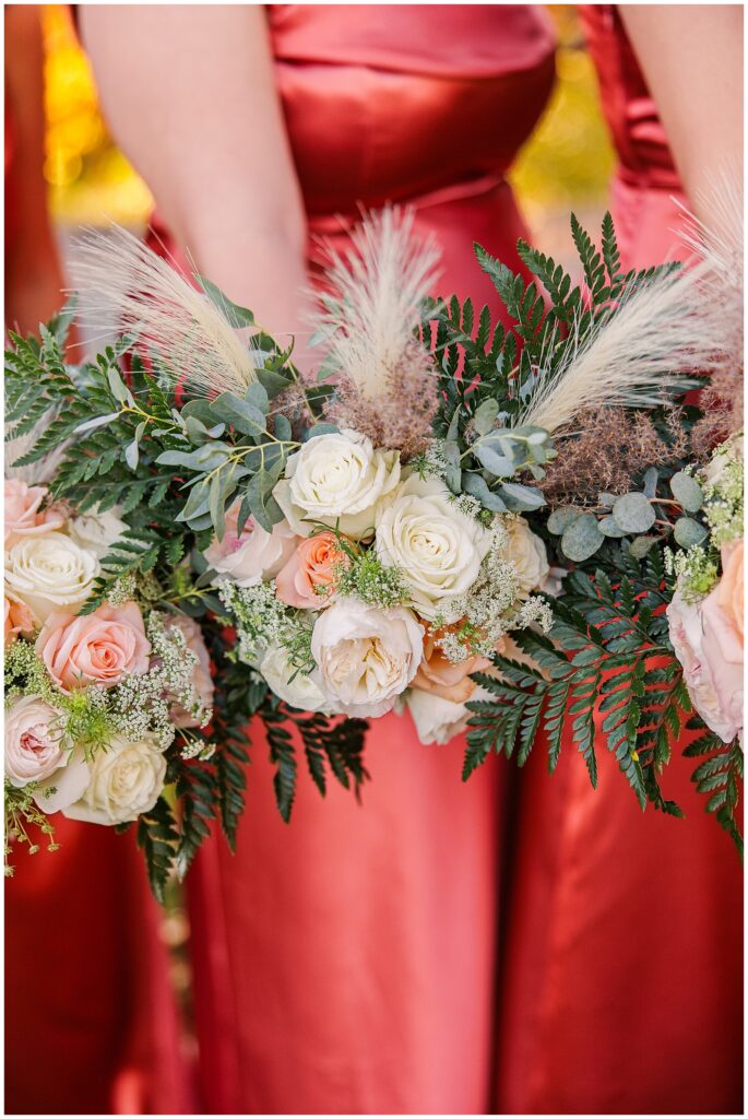 A close-up of bridesmaids holding matching bouquets of white and peach roses with greenery, pampas grass, and ferns. Their terracotta dresses contrast with the natural, autumn-inspired floral arrangements.