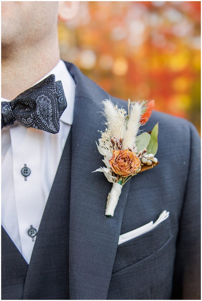 A groom at the Colonial Hotel wedding wears a dark suit with a black paisley bowtie and a boutonniere featuring dried florals, including a small orange rose, pampas grass, eucalyptus, and other fall accents.