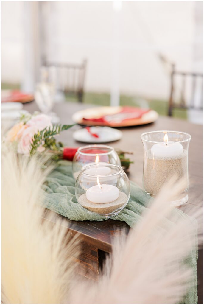 A fall-themed wedding table setup at the Colonial Hotel with candles in glass holders filled with sand and a soft green cloth draped down the center. In the background, place settings with red napkins and gold chargers are arranged on a wooden table.