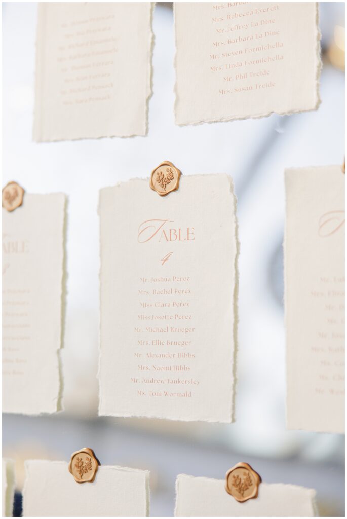 A seating chart display at the Colonial Hotel wedding featuring individual table assignments printed on textured paper with torn edges, each card sealed with a wax stamp featuring a floral design.