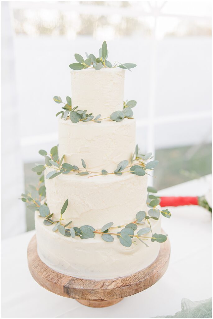 A simple three-tier wedding cake with smooth ivory frosting and eucalyptus sprigs around each layer. The cake sits on a wooden base, adding a natural touch to this fall wedding at the Colonial Hotel.