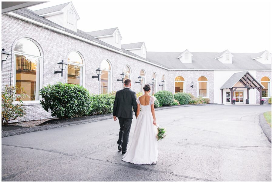 A bride and groom walk hand-in-hand along the driveway of the Colonial Hotel, with the bride holding her bouquet and wearing a white dress with a low back. The Colonial Hotel’s brick exterior and large arched windows provide a classic New England backdrop.