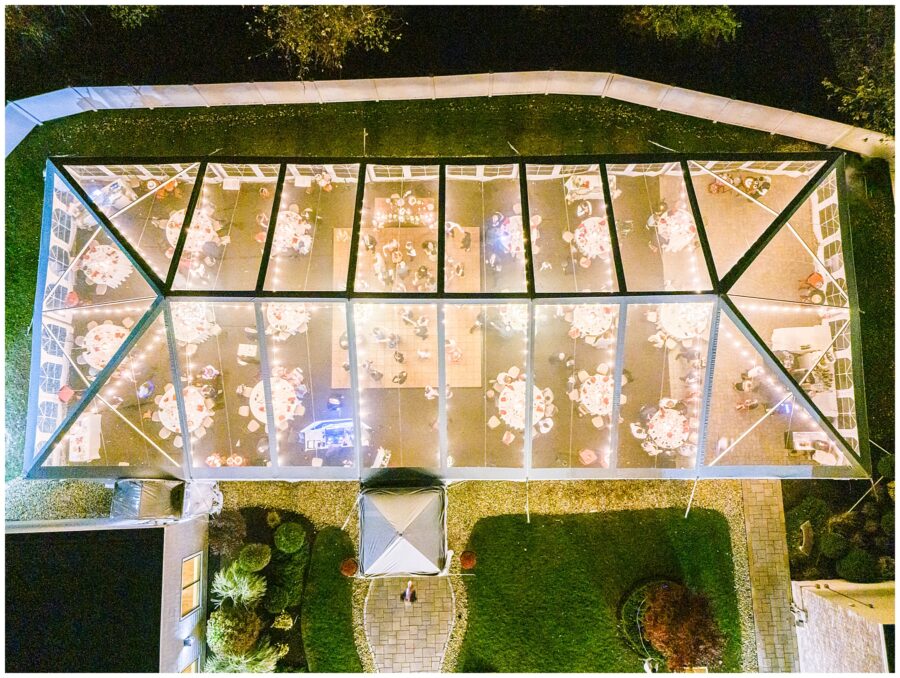 An overhead night drone shot of a large clear tent set up at the Colonial Hotel for a wedding reception. Inside the tent, tables with white tablecloths and red accents are arranged around a dance floor, with warm string lights creating a cozy ambiance.