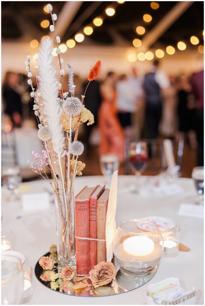 A fall-inspired wedding centerpiece at the Colonial Hotel, featuring vintage books, dried flowers, and soft candlelight. The table decor includes books with red and orange tones, a glass vase with dried florals in neutral colors, and sand-filled candles, creating a warm, vintage look against the softly lit background of the reception.