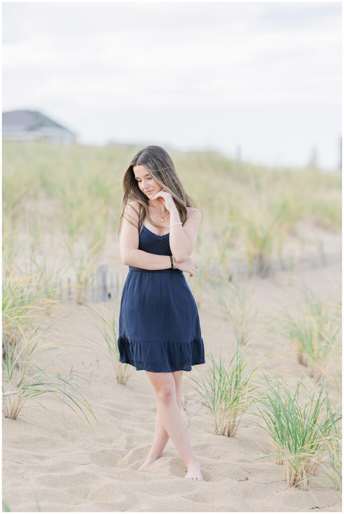 A young woman in a navy blue dress stands barefoot in the sand on a beach, surrounded by tall grasses. She looks down with a soft smile, lightly touching her face with one hand during a senior photo session in Newburyport, MA.