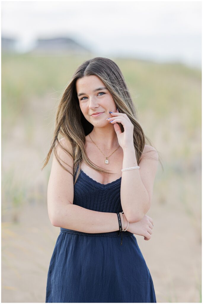 A close-up of the young woman in a navy dress, smiling gently and holding her hand near her face. Her long, straight hair falls around her shoulders, with the beach grass in the soft background, taken during her Newburyport, MA senior photo session.