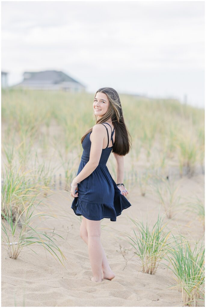 The young woman, barefoot and wearing a navy blue dress, turns back and smiles while walking through a grassy dune area on a private beach in Newburyport, MA, as part of her senior photo session.