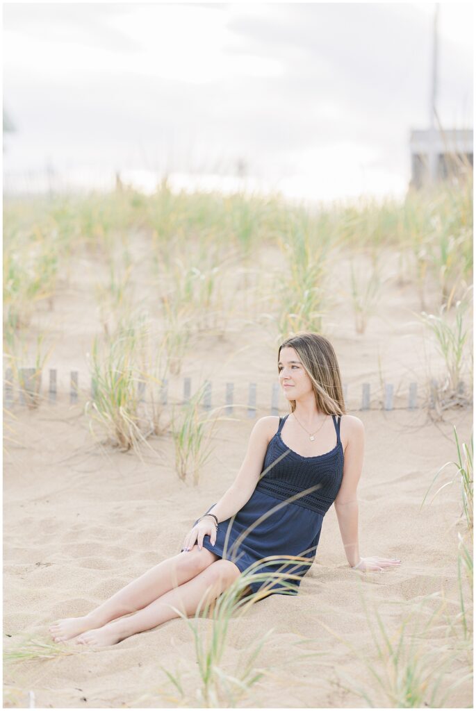 The young woman sits on the sandy beach, leaning back on her hands while looking off to the side. Her navy dress and the tall grass around her create a relaxed, coastal vibe during the senior photo session in Newburyport, MA.