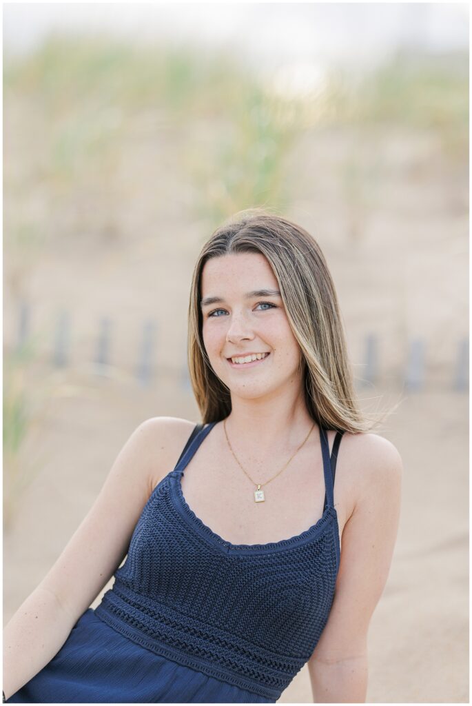 A close-up shot of the young woman sitting in the sand, wearing a navy blue dress and smiling slightly. Her long hair flows over her shoulders, with the beach grass softly blurred in the background.