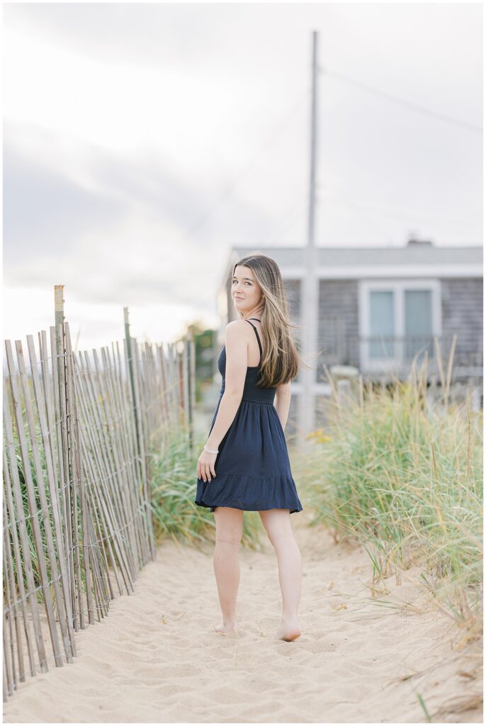 The young woman in a navy dress walks along a sand path lined with beach grass and a wooden fence. She glances back over her shoulder, with a beach house visible in the distance during her Newburyport, MA senior photo session.