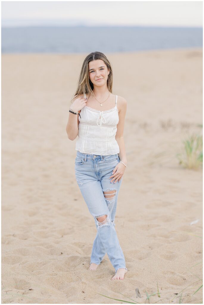 The young woman, now wearing a white sleeveless top and light blue ripped jeans, stands barefoot on the sandy beach during her senior photo session in Newburyport, MA, with the ocean in the background.