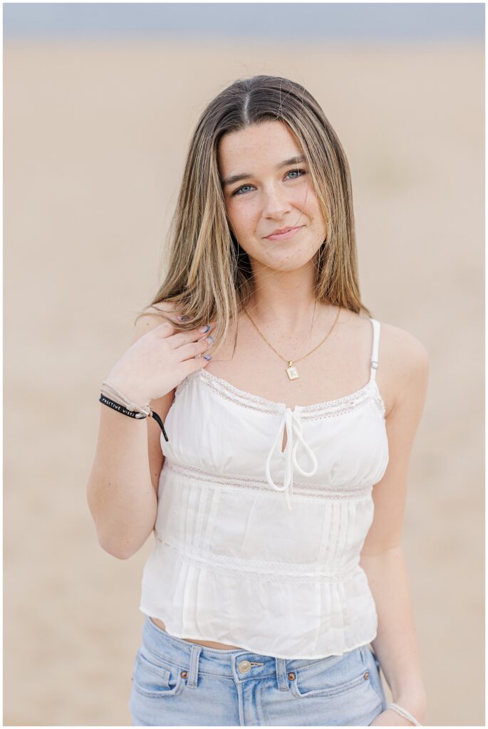 A close-up of the young woman in a white top and jeans, smiling softly. She stands against the backdrop of the beach with her hand gently touching her hair, taken during her senior photo session on a private beach in Newburyport, MA.