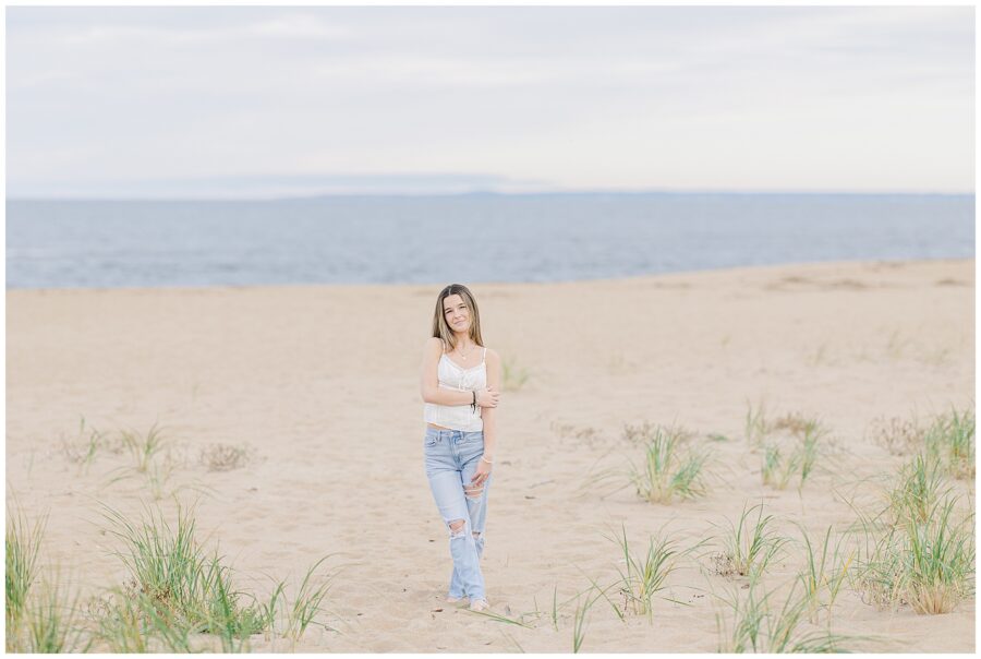 The young woman walks along the sandy beach in Newburyport, MA, barefoot and dressed in a white top and jeans. The ocean is in the background, and scattered beach grass adds to the natural, relaxed setting of her senior photo session.