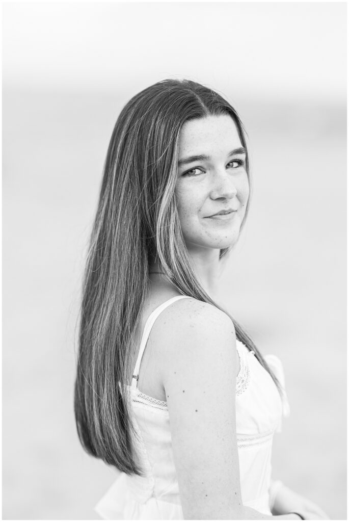 A black-and-white portrait of a young woman looking over her shoulder, with long hair and a slight smile. She wears a white top during a senior photo session on a private beach in Newburyport, MA.