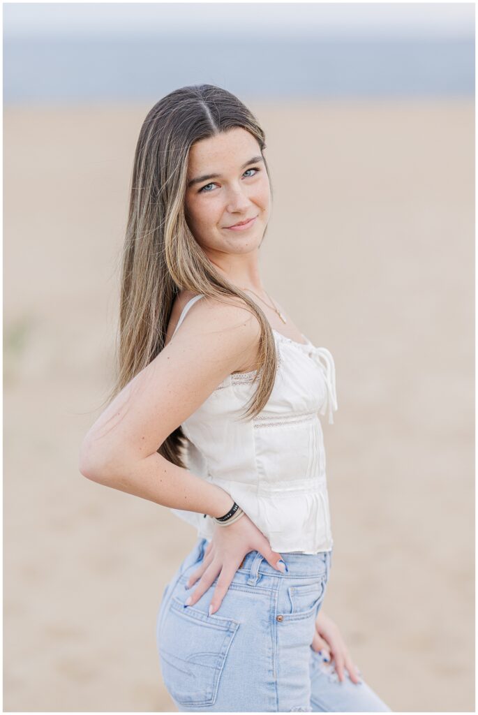 The young woman in a white top and ripped jeans poses with one hand on her hip, smiling softly. The sandy beach of Newburyport, MA, serves as the backdrop for this senior photo session.