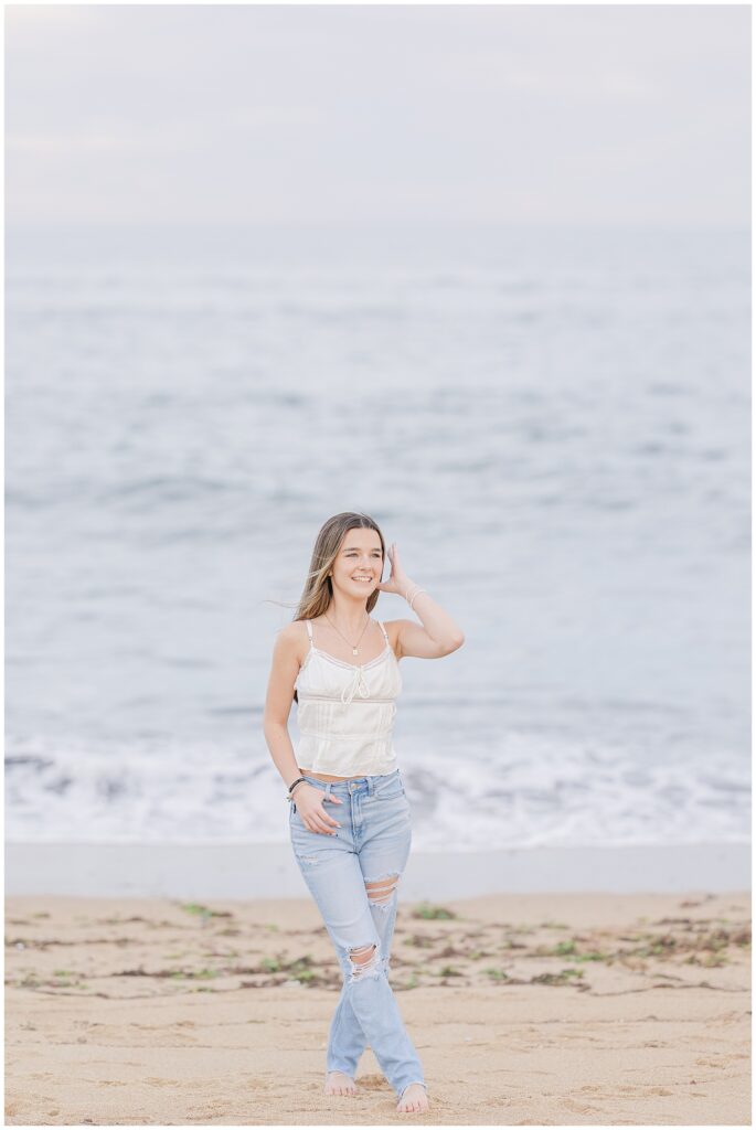 A candid shot of the young woman walking along the shore, smiling and lightly touching her hair, with waves crashing in the background. She is barefoot, wearing a white top and light blue ripped jeans during her senior photo session in Newburyport, MA.