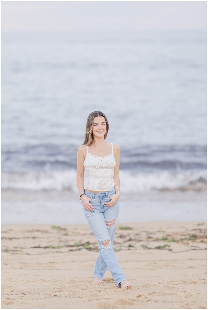 The young woman stands near the water’s edge, smiling gently as the ocean waves roll in behind her. She wears a white top and light blue jeans for her senior photos at a private beach in Newburyport, MA.