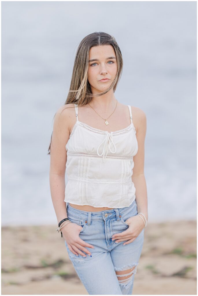 A close-up of the young woman standing on the beach, with her hands in her pockets and a thoughtful expression. She wears a white top and ripped jeans during her senior photo session in Newburyport, MA.