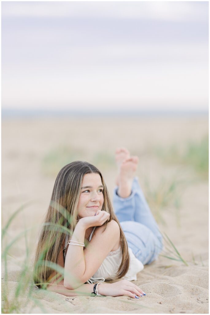 The young woman lies on her stomach in the sand, propped up on her elbows with a relaxed smile. She is barefoot and wearing a white top and jeans, photographed during her senior session at a private beach in Newburyport, MA.