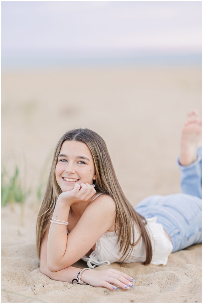 A smiling shot of the young woman lying on her stomach in the sand, with her head resting on her hand. She wears a white top and light blue jeans for her senior photo session on a Newburyport, MA beach.