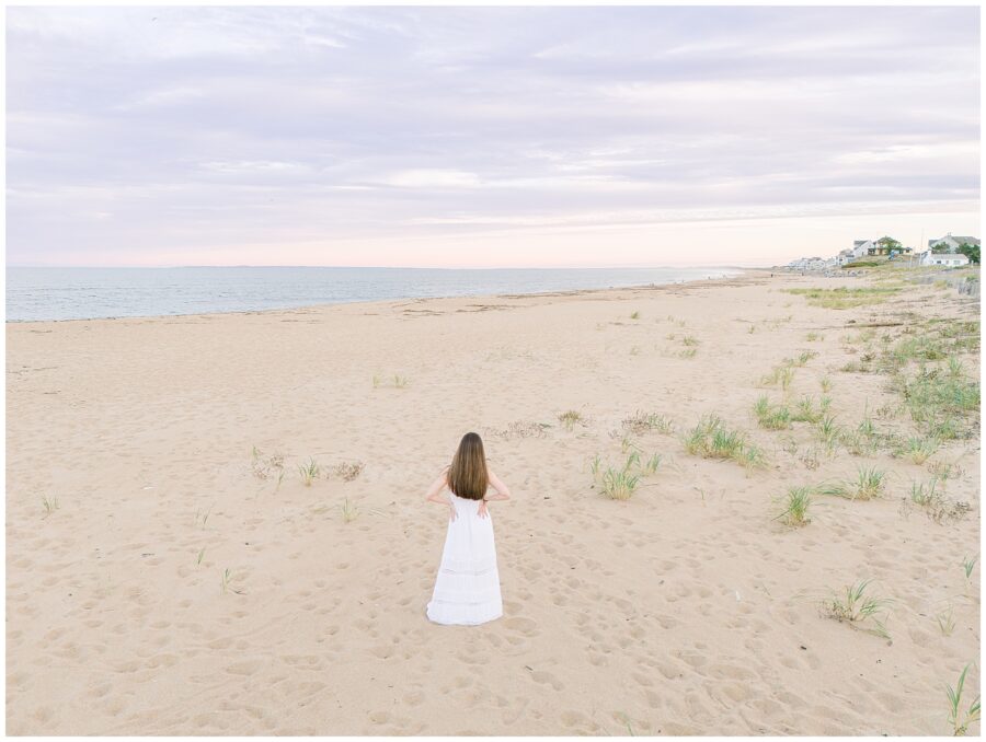 A wide shot of the young woman walking away from the camera along the vast sandy beach, wearing a white dress. The shoreline extends in the distance, showcasing the natural beauty of the private beach in Newburyport, MA.