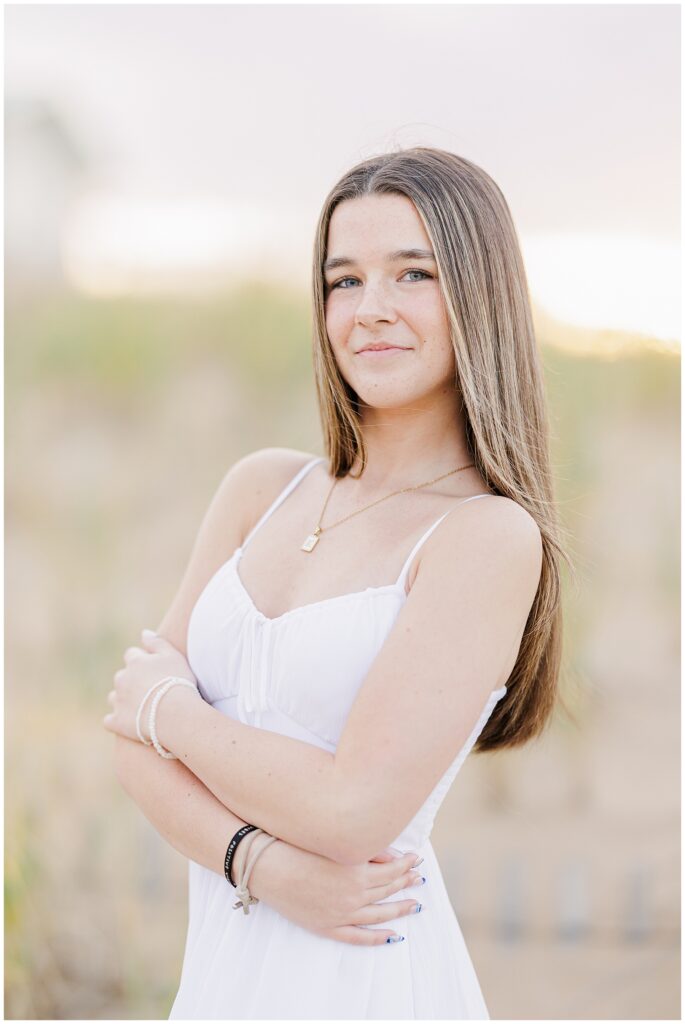 A close-up of the young woman standing with her arms crossed, wearing a white dress. She smiles softly at the camera, capturing the serene atmosphere of her senior photo session at a private beach in Newburyport, MA.