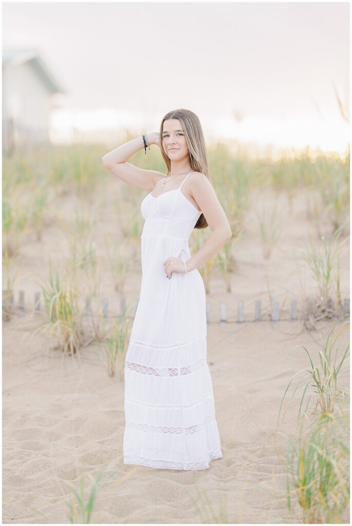 A young woman poses confidently in a flowing white dress, hand on her hip, with beach grass and soft sunset light in the background during her senior photo session on a private beach in Newburyport, MA.