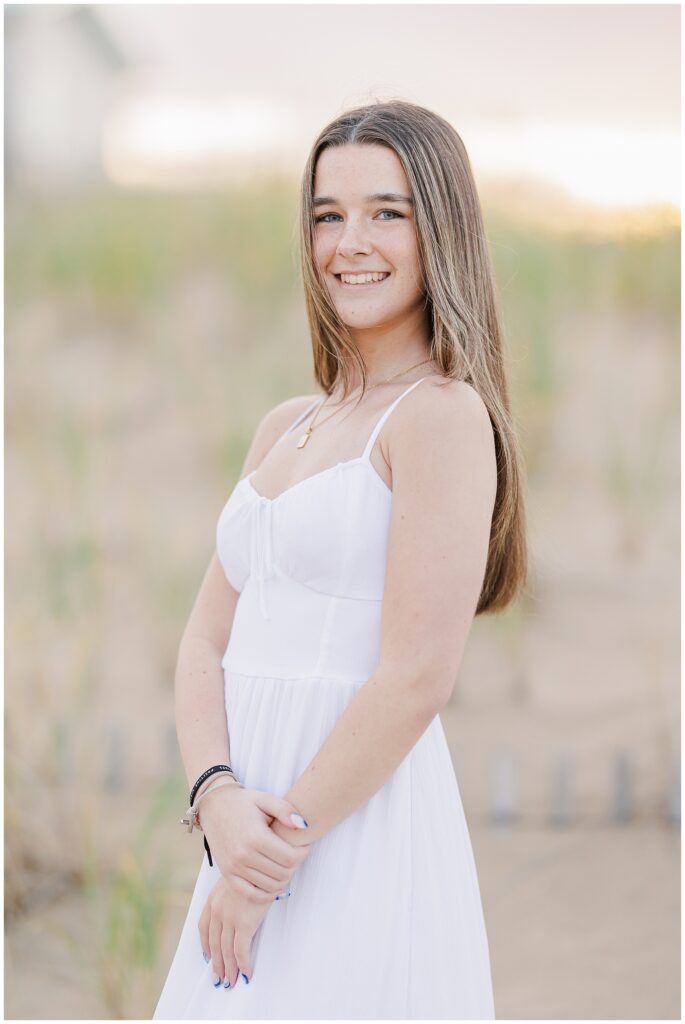 The young woman smiles with her arms crossed, wearing a white dress. The natural beach setting of Newburyport, MA, frames the background for her senior photos.