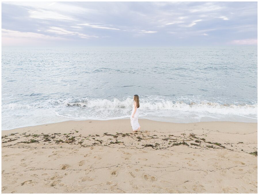 A wide shot of the young woman walking along the shoreline, with gentle waves crashing at her feet. She wears a white dress during her senior photo session at a private beach in Newburyport, MA.