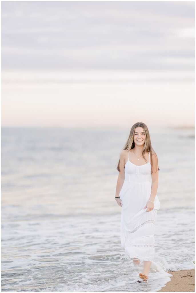 The young woman walks barefoot in the water, smiling as the waves lap at her feet. She is dressed in a white flowing dress, capturing a serene moment during her Newburyport, MA senior photo session.