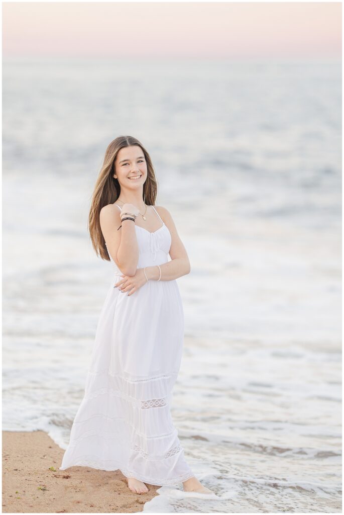 The young woman stands barefoot at the water’s edge, smiling softly. She wears a white dress as the waves swirl around her feet during her senior photo session in Newburyport, MA.