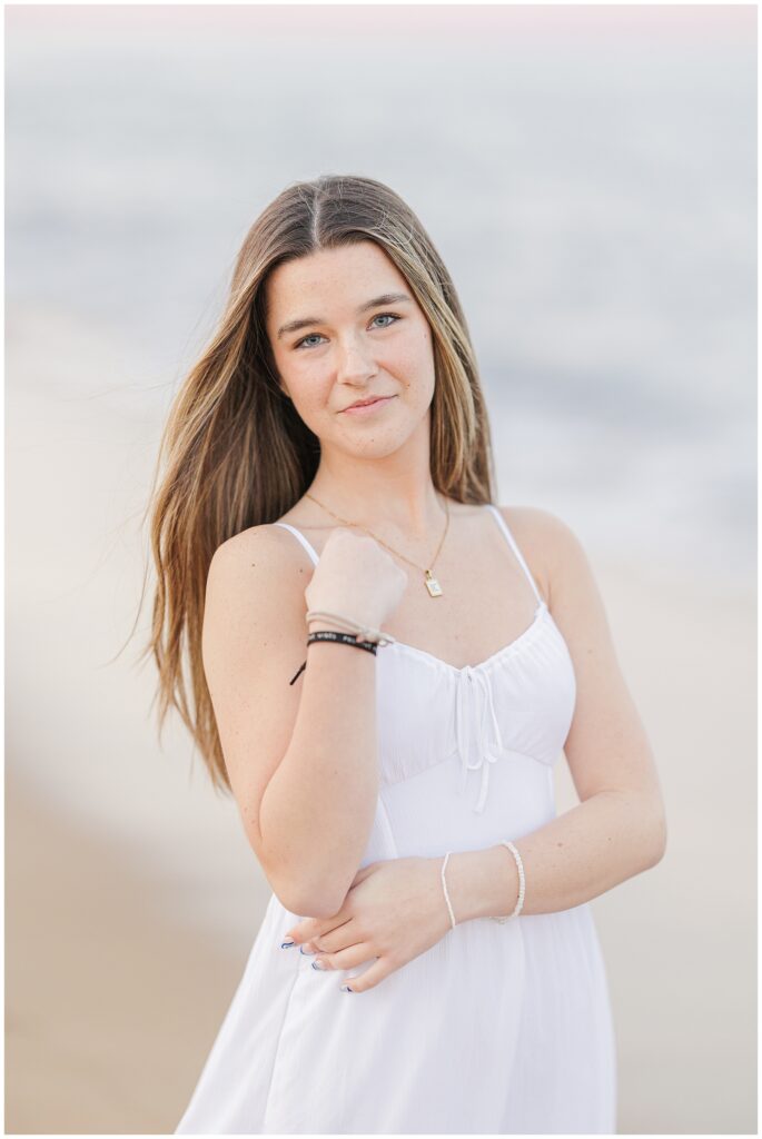 A close-up of the young woman standing on the beach, smiling with her hair gently blowing in the breeze. She wears a white dress for her senior photos at a private beach in Newburyport, MA.