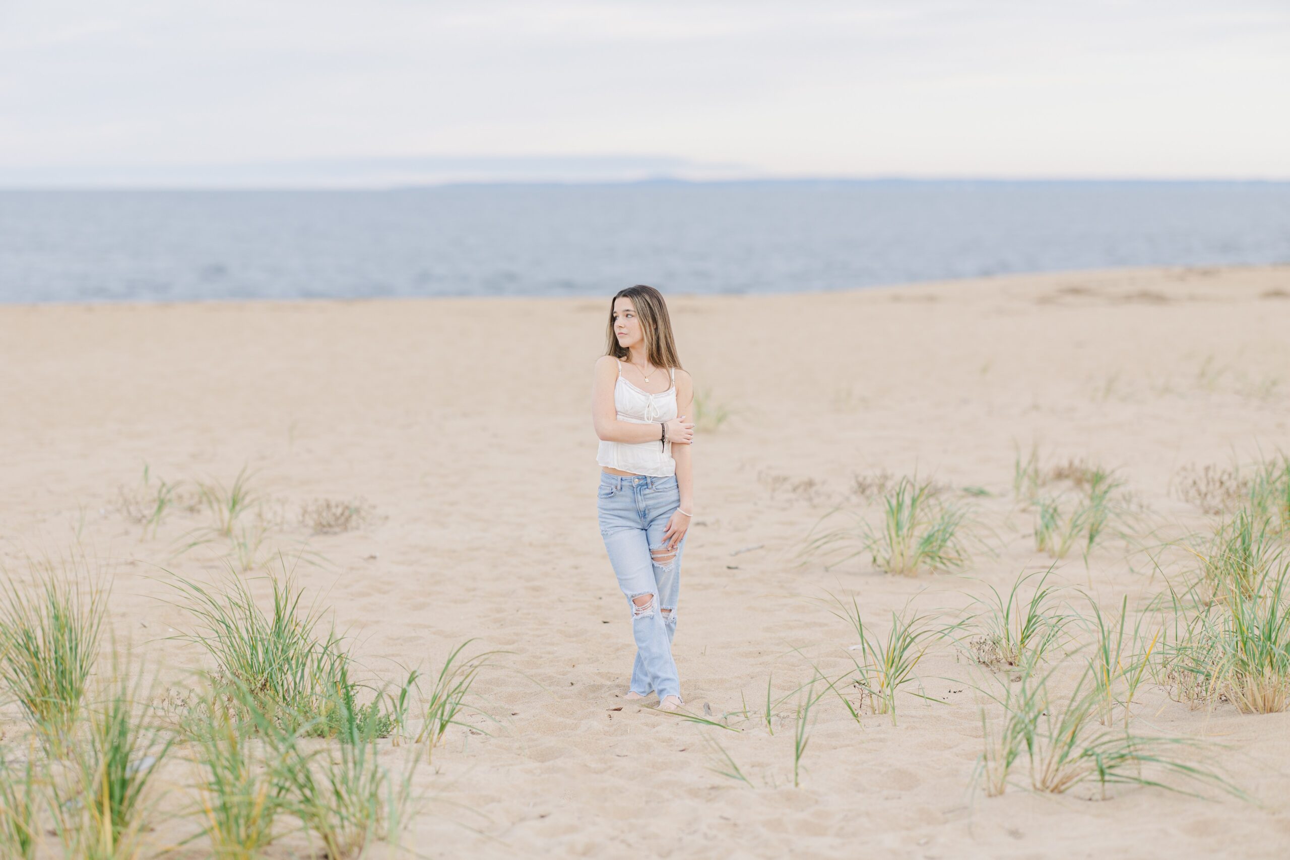 A young woman stands barefoot on a sandy beach in Newburyport, MA, wearing a white sleeveless top and ripped jeans. She poses with one arm crossed and looks off to the side, surrounded by scattered beach grass with the ocean in the background. This image was captured during a senior photo session by a Newburyport photographer.