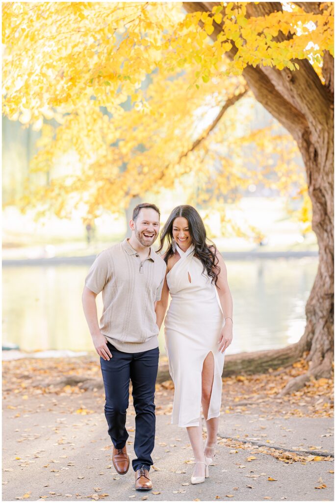 A couple walking arm-in-arm along a path at Boston Public Garden, surrounded by golden fall foliage, with a calm pond in the background. The woman wears a white dress with a thigh slit, and the man is dressed in a beige shirt and dark pants. Perfect for showcasing Boston engagement pictures.