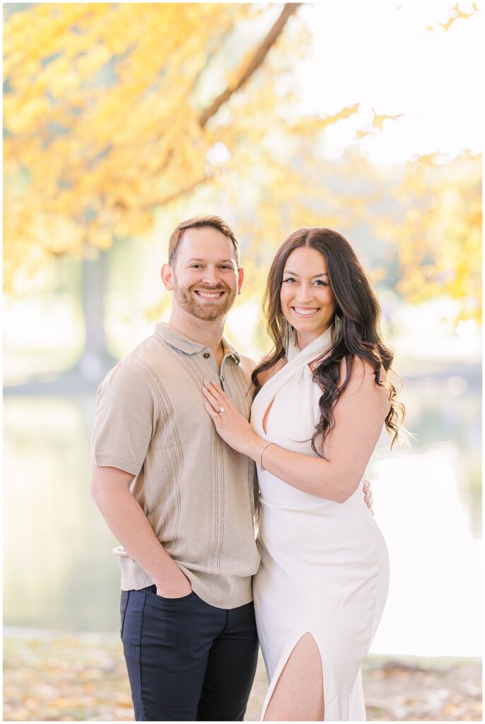 The couple standing close together at Boston Public Garden, smiling directly at the camera. The woman has her hand on the man’s chest, displaying her engagement ring. Bright autumn leaves frame the scene, adding warmth to these Boston engagement pictures.