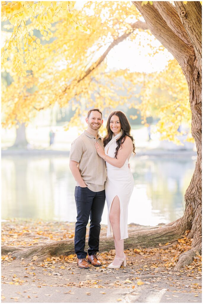 A wide shot of the couple standing near a tree with vibrant yellow leaves at Boston Public Garden. The man has his hand in his pocket, while the woman stands beside him in a white dress, creating a classic moment for Boston engagement pictures.