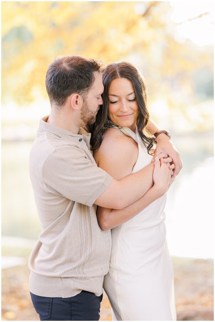 The man embracing the woman from behind, his arms wrapped around her. The woman looks down with a soft smile. Yellow autumn leaves and the pond at Boston Public Garden form a romantic backdrop.