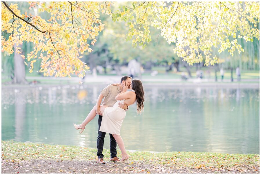 A playful scene where the man lifts the woman off the ground near the pond in Boston Public Garden. The woman has her arms around his neck and her legs slightly bent, as vibrant fall leaves dangle above them.