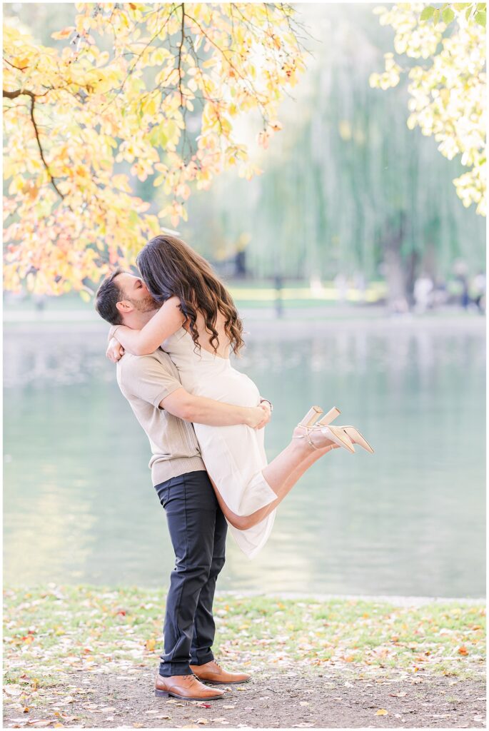 The couple shares a kiss as the man lifts the woman into the air near the pond in Boston Public Garden. The woman’s beige heels and flowing white dress are captured mid-motion, with autumn foliage framing the image.