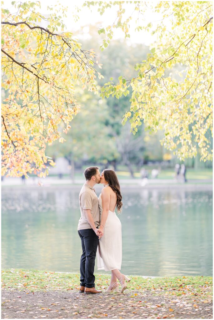 The couple holds hands and shares a kiss at Boston Public Garden, standing near the edge of the pond. The golden leaves overhead create a dreamy atmosphere, ideal for Boston engagement pictures.