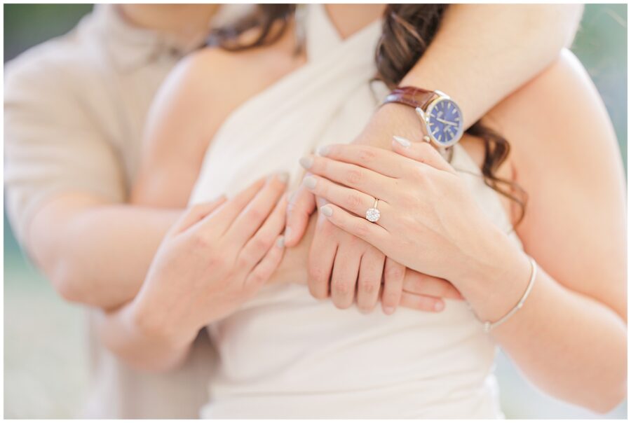 A close-up of the couple’s hands wrapped around each other. The focus is on the woman’s engagement ring and a man’s watch, symbolizing their commitment. The backdrop is softly blurred.